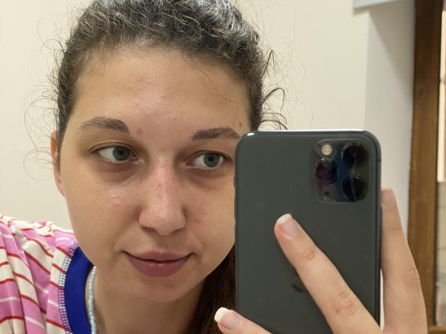 Sanella with messy brown hair, holding a black iPhone, gray eyes, slightly flushed face, pink-striped shirt, subtle smirk, and small zit on the nose, in a selfie.