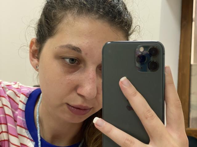 Sanella with brown curly hair, blue eyes, and pink striped shirt, holding a grey iPhone, taking a selfie, showing a slight pout, with a small nose piercing.