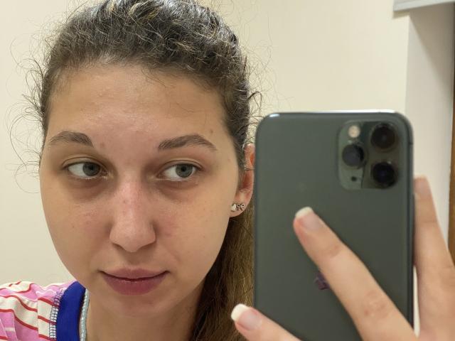 Sanella has a dirty face with small tits, curly brown hair in a ponytail, and is taking a selfie with a grey iPhone, showing a neutral expression.