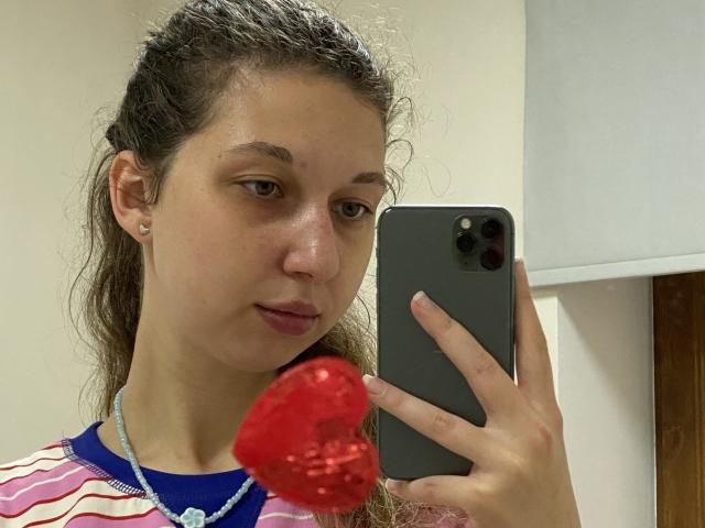 Sanella with wet, curly brown hair, wearing a striped shirt, holding a red, textured ball in front of her face, taking a selfie with a gray iPhone.