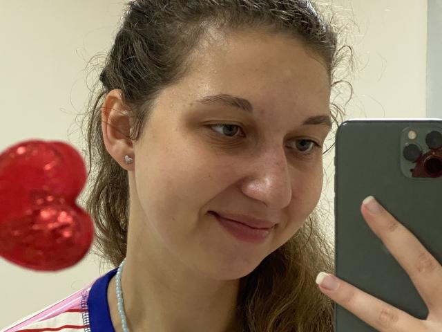 Sanella's face in a selfie, brown curly hair, light skin, small earrings, subtle smile, pink shirt, holding a gray phone, red heart-shaped balloon on the left.