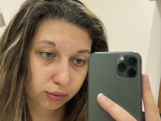 Sanella with wavy brown hair, pale skin, green eyes, and a slight pimple on her nose, holding a green iPhone, taking a selfie. Her pink lips are slightly parted.