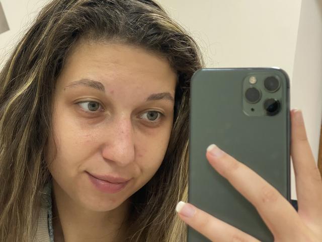 Sanella with long brown hair, green eyes, slight smile, small zit on nose, holding gray iPhone, wearing nothing visible, looking into camera, showing pinkish lips, and pale skin.