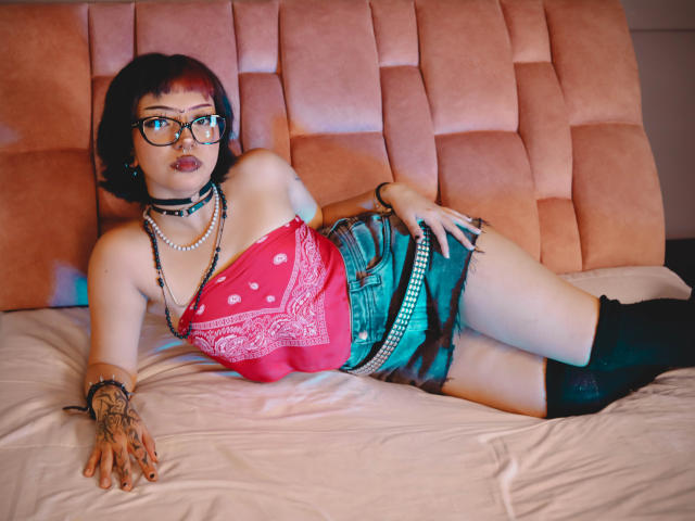 JadeMilleer with red-tipped black hair, glasses, choker, pink bandana top, green camo shorts, black thigh-highs, and tattoos, laying on a pink tufted bed, showing her tits.