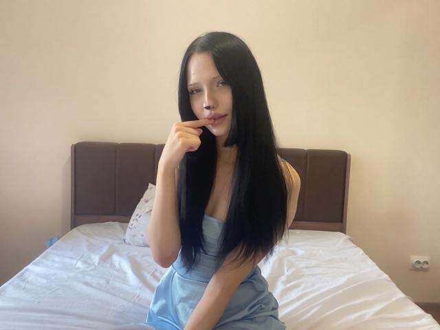 CherelleGess with long black hair, wearing a strapless blue dress, sitting on a bed, finger touching her lip, looking at the camera, with a subtle smile.