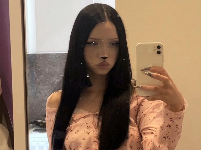 CherelleGess with long black hair, wearing a pink off-shoulder top, holding a white phone, looking in a mirror, showing her tits and small ass, with fake eyelashes.