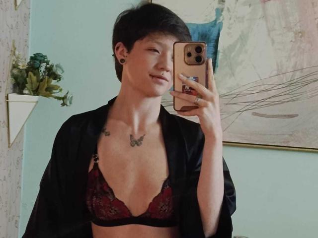 LissyLuxe with short dark hair, wearing a black robe and red lace bra, holding a phone, showing small tits with a butterfly tattoo on her chest.