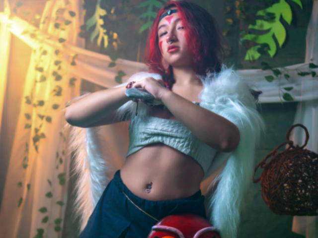 Kanela Miller, con cabello rojo, alas blancas con plumas y una diadema roja, vestida con un top corto gris que muestra su abdomen tonificado y su ombligo perforado, sosteniendo un paño gris..