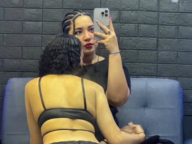 CharlotteTiffany with braided hair, red lipstick, and black top, taking a selfie while sitting on a blue couch. Another woman with curly hair, black bra, and tan skin.