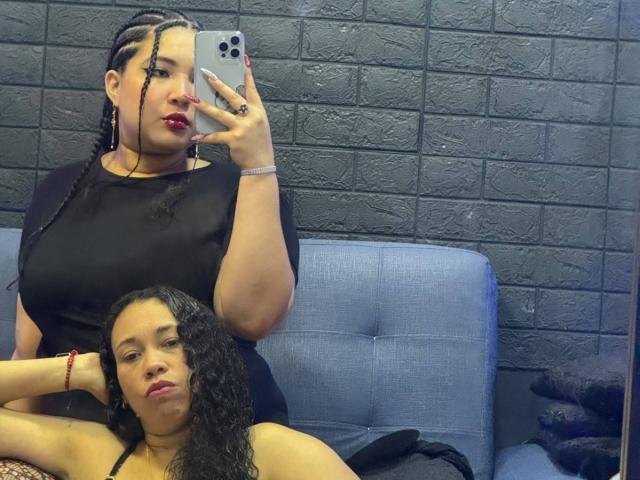 CharlotteTiffany with big boobs, wearing a black top, holding a phone, taking a mirror selfie. She's sitting next to a girl with curly hair, on a blue couch.