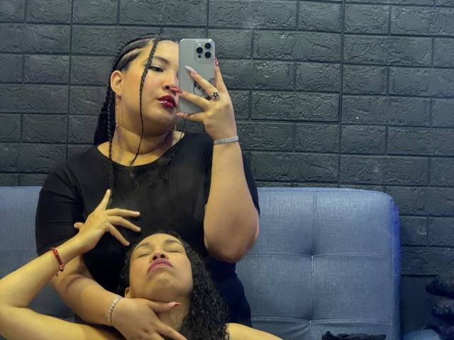 CharlotteTiffany with braids, black shirt, red lipstick, holding phone, taking selfie. Her hand's on another woman's neck, who's lying back, eyes closed, black hair, no shirt.