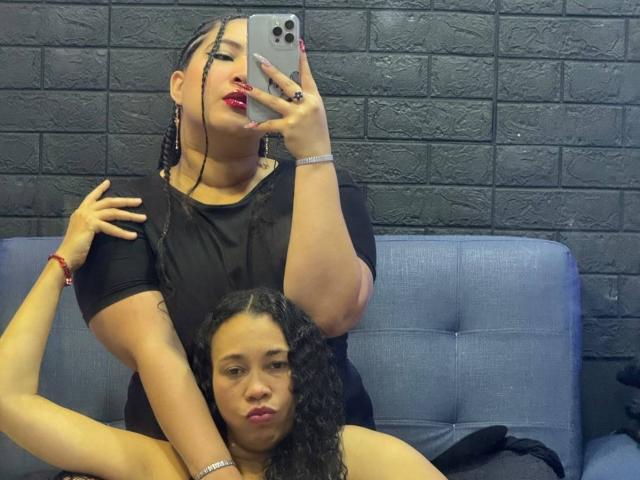 CharlotteTiffany with braided pigtails, red lipstick, black shirt, holding phone, arm around curly-haired woman's shoulder, both sitting on blue couch, black brick wall in background.