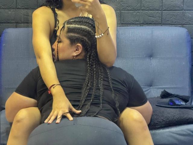 CharlotteTiffany with braided hair, kissing a black-shirted dude's neck, her hand on his ass, sitting on a blue sofa, her tits visible, holding a phone.