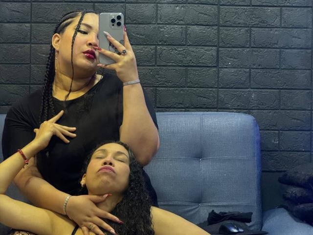 CharlotteTiffany with long black braids and red lipstick, holding phone, sits on blue chair, her hand on smaller CharlotteTiffany's neck, who has curly hair, both in black shirts.