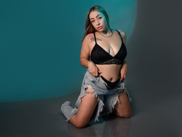 CharlotRey, ajoelhada, vestindo um sutiã de renda preta, jeans rasgados, puxando a calcinha preta para baixo, mostrando a vulva, tatuagens no ombro, luz azul-esverdeada, expressão séria, cabelo castanho longo, batom rosa..
