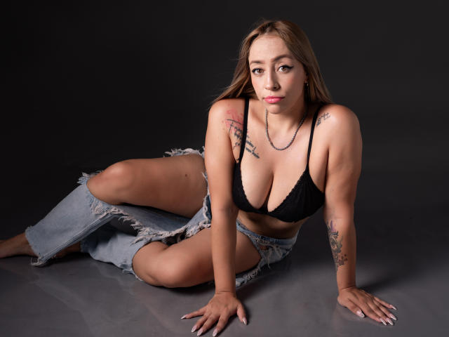 CharlotRey with tits in a black bra, ripped jeans, tattoos, and a necklace, sitting on the floor, looking at the camera, with a neutral expression.