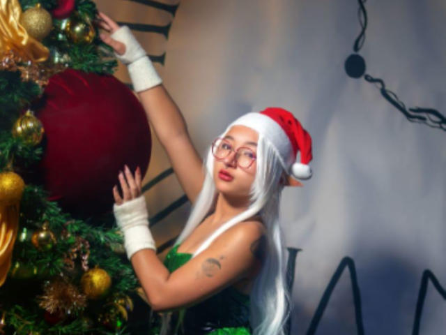 KanelaMiller mit weißen Elfenohren, einem roten Weihnachtsmannhut, Brille, einem grünen Kleid, weißen Armbändern, die eine Weihnachtsbaum mit goldenen und roten Ornamenten berührt, und die ein Tattoo auf ihrem Arm zeigt..