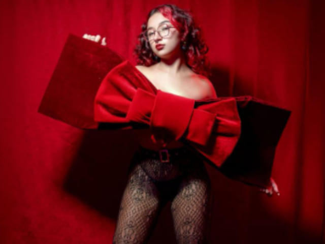 KanelaMiller con cabello rizado rojo, gafas, blusa roja de terciopelo con un hombro descubierto, pantalones negros de encaje, sosteniendo la parte superior, fondo rojo. Los senos son ligeramente visibles debajo de la blusa. Pose sexy y segura..