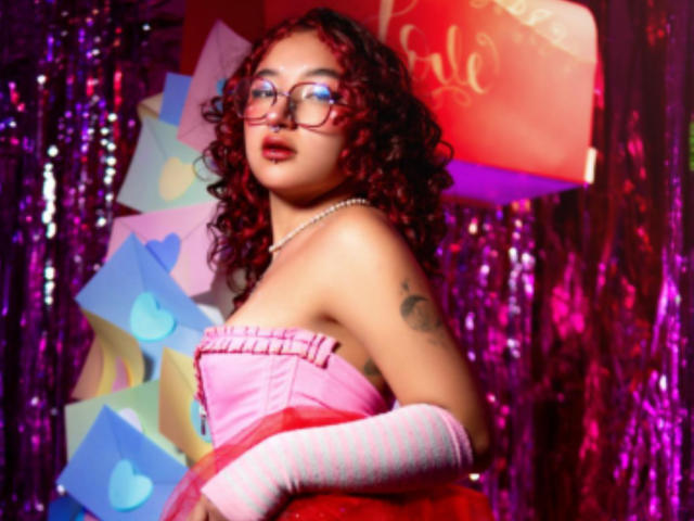 Kanela Miller mit lockigem, rotem Haar, die eine Brille trägt, ein pinkes, trägerloses Kleid und pinke, gestreifte Handschuhe, zeigt ein Tattoo auf ihrem rechten Arm und trägt eine Perlenkette..