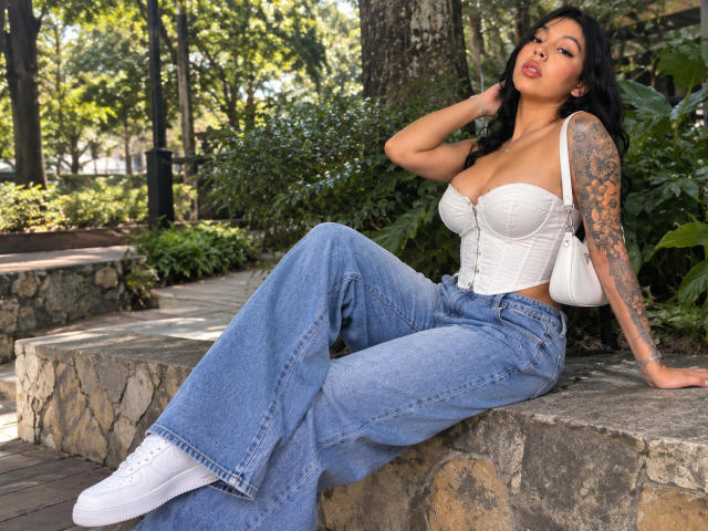 NenaBlack with tattoos, white strapless top, and blue flares, sitting on stone, hand in hair, showing her big tits and white sneakers, in a sunny park.