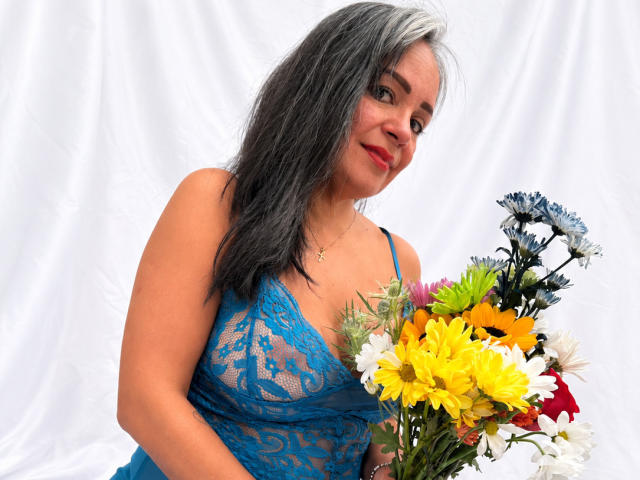 LailaCastillo with gray and white hair, blue lace dress, holding bouquet of yellow sunflowers, white daisies, and blue flowers, smiling at camera, showing medium tits.