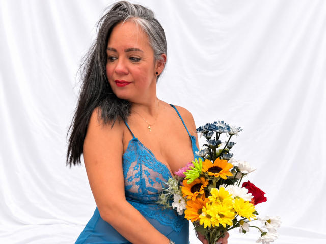 LailaCastillo with gray and black hair, wearing blue lace lingerie, holding a bouquet of sunflowers, yellow daisies, and red roses, smiling slightly, red lipstick, white background.