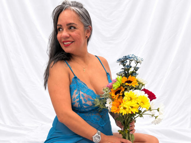 LailaCastillo with gray hair, blue lace dress, holding bouquet of sunflowers, yellow daisies, blue flowers, sitting, smiling, red lipstick, wearing watch, white background.