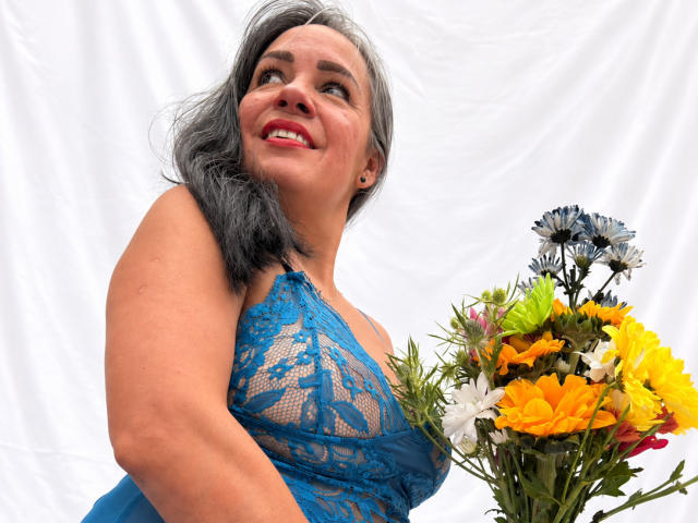 LailaCastillo with gray hair, blue lace dress, holding colorful bouquet, smiling up, showing her tits, holding flowers in right hand, white background, red lipstick.
