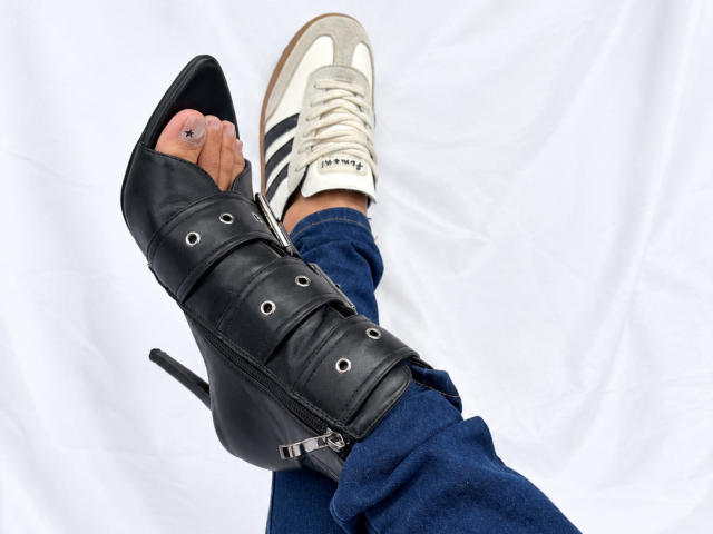 LailaCastillo's foot in a black, high-heeled, strappy leather boot with metal rings, next to a white sneaker with black stripes, blue jeans, and a visible toe ring.
