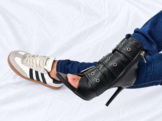 LailaCastillo's foot in a white Adidas sneaker with black stripes, while the other foot is in a black high-heeled boot with multiple straps, wearing blue jeans.