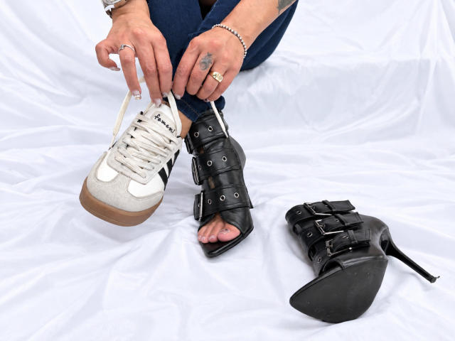 LailaCastillo's hands tying white sneaker laces, wearing black strappy high heel on foot, blue jeans, white sheet, tattoos visible, ring on finger, second black heel on ground.