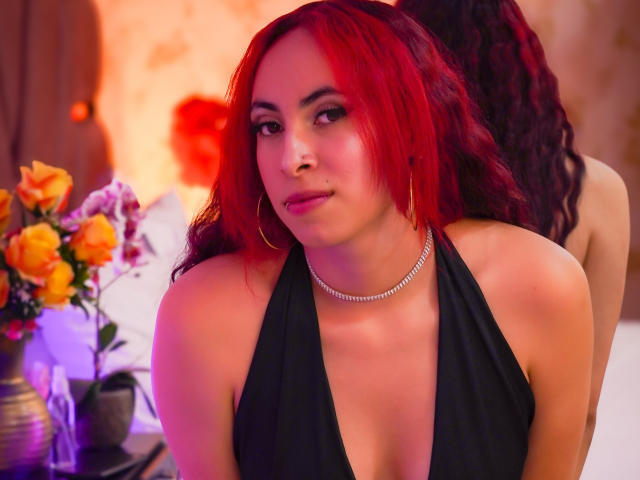 Anya Carter, con cabello rojo, un top de tirantes negros y un collar, mostrando sus senos, está de pie con un trasero desnudo en el fondo. Lleva aros dorados y parece ligeramente aburrida..