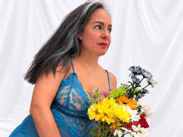 LailaCastillo with grayish-black hair, wearing blue lace dress, holding colorful bouquet, looking to the side, red lipstick, slightly chubby, small tits, white fabric background.