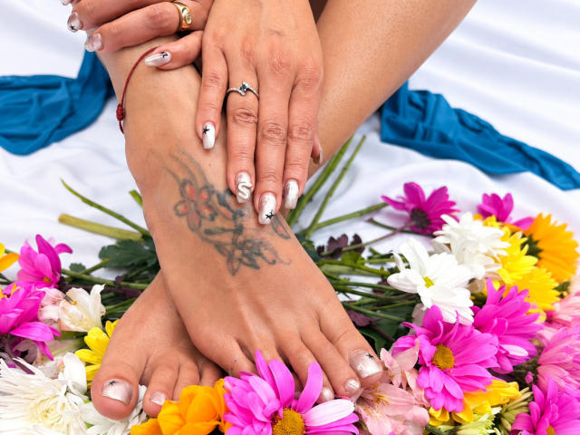 LailaCastillo's feet with a tattoo on her left sole, hands with rings and floral nail art, resting on colorful flowers. Her toes are painted white with pink tips.