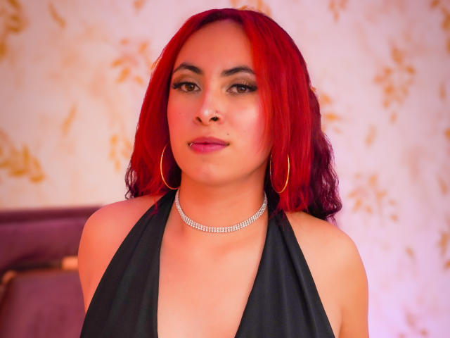 AnyaCarter with red hair, hoop earrings, and a diamond choker, wearing a black halter top, showing her tits and a subtle nose piercing, looking confident.