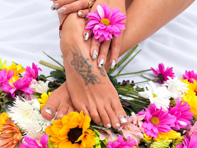 LailaCastillo's feet with a tattoo, pink flower, and silver nail polish, resting on colorful flowers, including pink, yellow, and white daisies. Her toes are painted metallic silver.