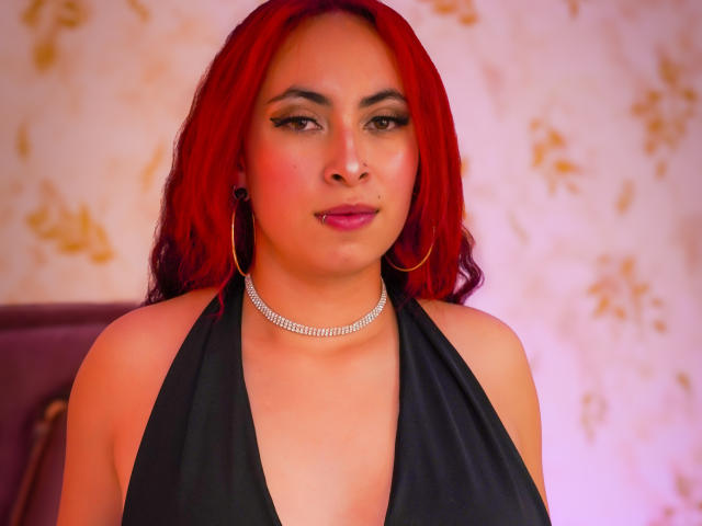 Anya Carter, met rood haar, draagt een zwarte haltertop, een diamanten ketting en oorringen, heeft volle lippen en een neusring, en kijkt recht in de camera..