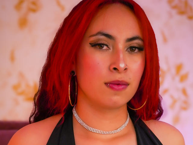 Anya Carter, met rood haar, oorringen, een diamanten ketting, roze lippenstift en een zwarte haltertop, toont een zelfverzekerde uitdrukking, waarbij haar borsten licht zichtbaar zijn. Haar make-up is opvallend en aantrekkelijk..
