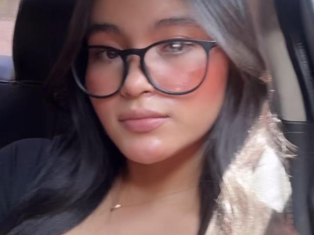 MiiaPalmer with big black glasses, pinkish-red cheeks, and long black-brown hair, sitting in a car, wearing a black top, and a small necklace, looking slightly to the side.