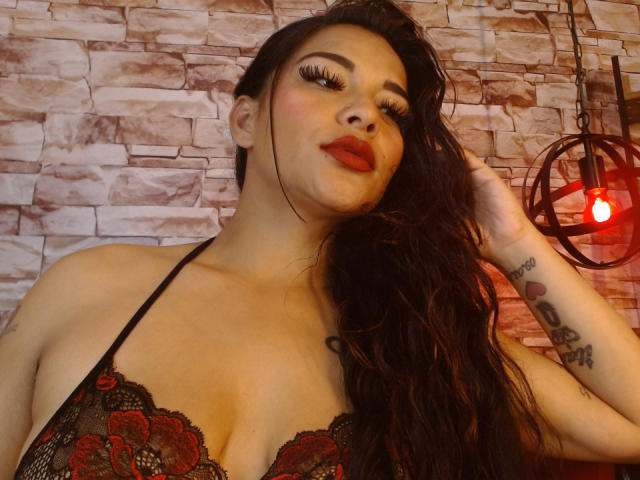 MiaBellucci with long, wavy dark hair, red lipstick, and heavy eyelashes, wearing a lace black and red bra, showing her tits, tattoos on right arm, and a red light.
