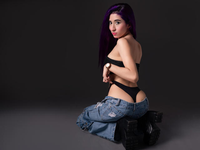JuliaJobs with purple hair, black strapless top, black thong, and blue jeans, kneeling, showing her ass, wearing black boots, and a watch, looking back.
