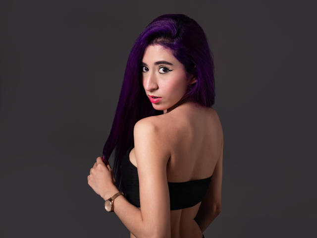 JuliaJobs with purple hair, black strapless top, looking over her shoulder, holding hair, pink lipstick, tan skin, small breasts, bracelet on her right wrist, dark gray background.