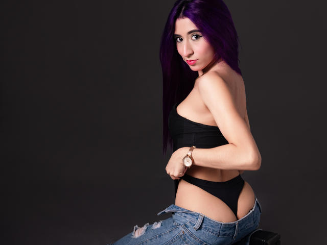 JuliaJobs with purple hair, black strapless top, black thong, and unbuttoned jeans, showing her ass, looking over her shoulder with a smile, wearing a gold watch.