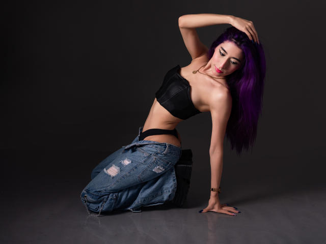 JuliaJobs with purple hair, black strapless bra, black panties, and blue jeans, kneeling, hand on head, showing her tits and ass, dark background, studio shot.
