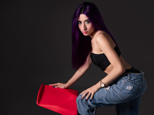 JuliaJobs with purple hair, black strapless top, and blue jeans, leaning on a red chair, wearing a watch, looking at the camera, dark background.