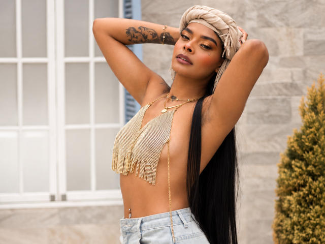EmmaEbony with long black hair, wearing a gold chain top that barely covers her small, bare tits, arms raised, beige headwrap, tattoo on left arm, standing outside.