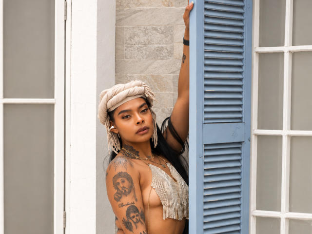 EmmaEbony with tattoos, dark skin, wearing a beige headwrap, blue shutters, and a mesh top, leaning on a window, looking at the camera, showing her small tits.