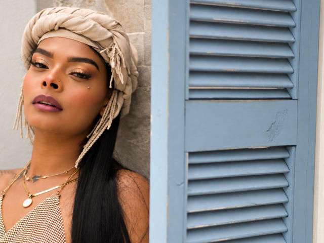 EmmaEbony with dark skin, long black hair, wearing a beige headwrap, gold mesh top, and jewelry, standing next to blue shutter, looking down with purple lipstick.