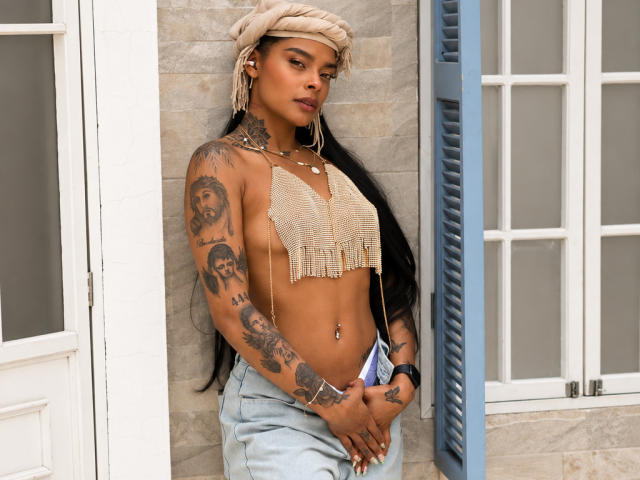 EmmaEbony with long black hair, tan skin, tattoos on arms and chest, wearing a beige headwrap, mesh top, and light blue jeans, standing against a building with blue shutters.