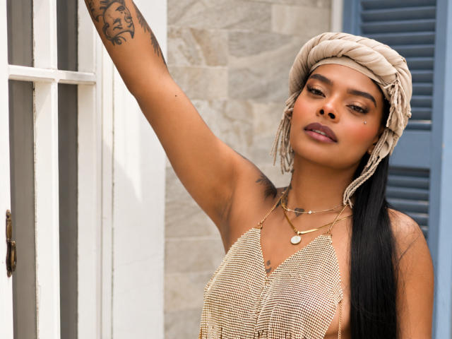 EmmaEbony with tan skin, dark brown hair in a ponytail, wearing a beige headwrap, gold necklace, and a striped top, raising her tattooed arm, showing off her tits.
